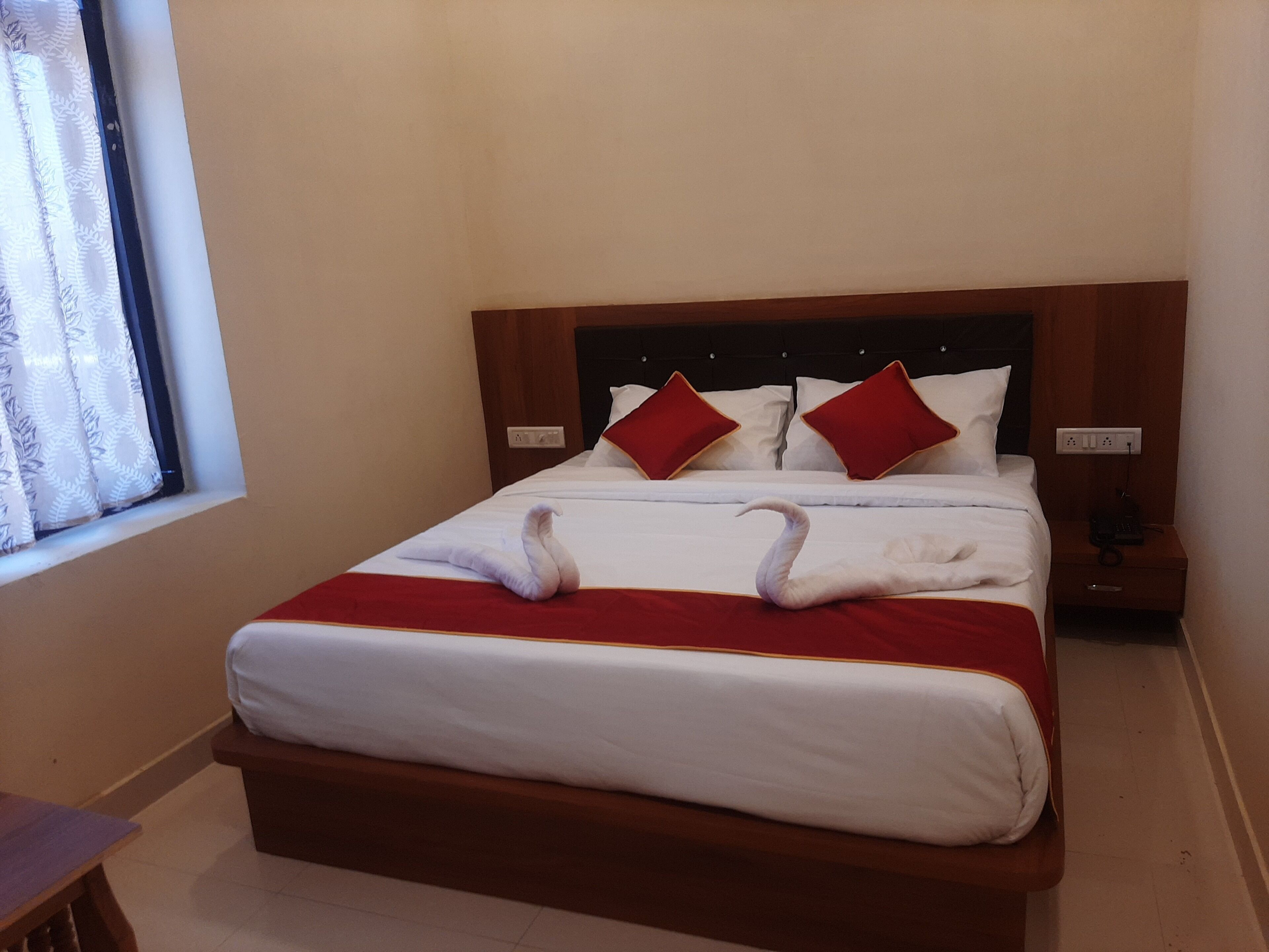 A neatly made bed with a red blanket and two red pillows, a wooden headboard, a bedside table with a phone, and a window with curtains.