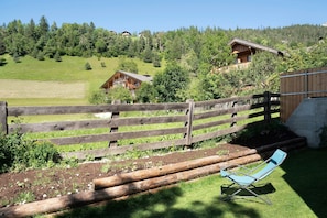 A wooden fence, a blue lounge chair, and a house with a balcony in a green, hilly landscape.