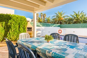 Un área de comedor al aire libre techada con una mesa lista para dos personas, rodeada de sillas, una planta en maceta y vistas a palmeras y un edificio.