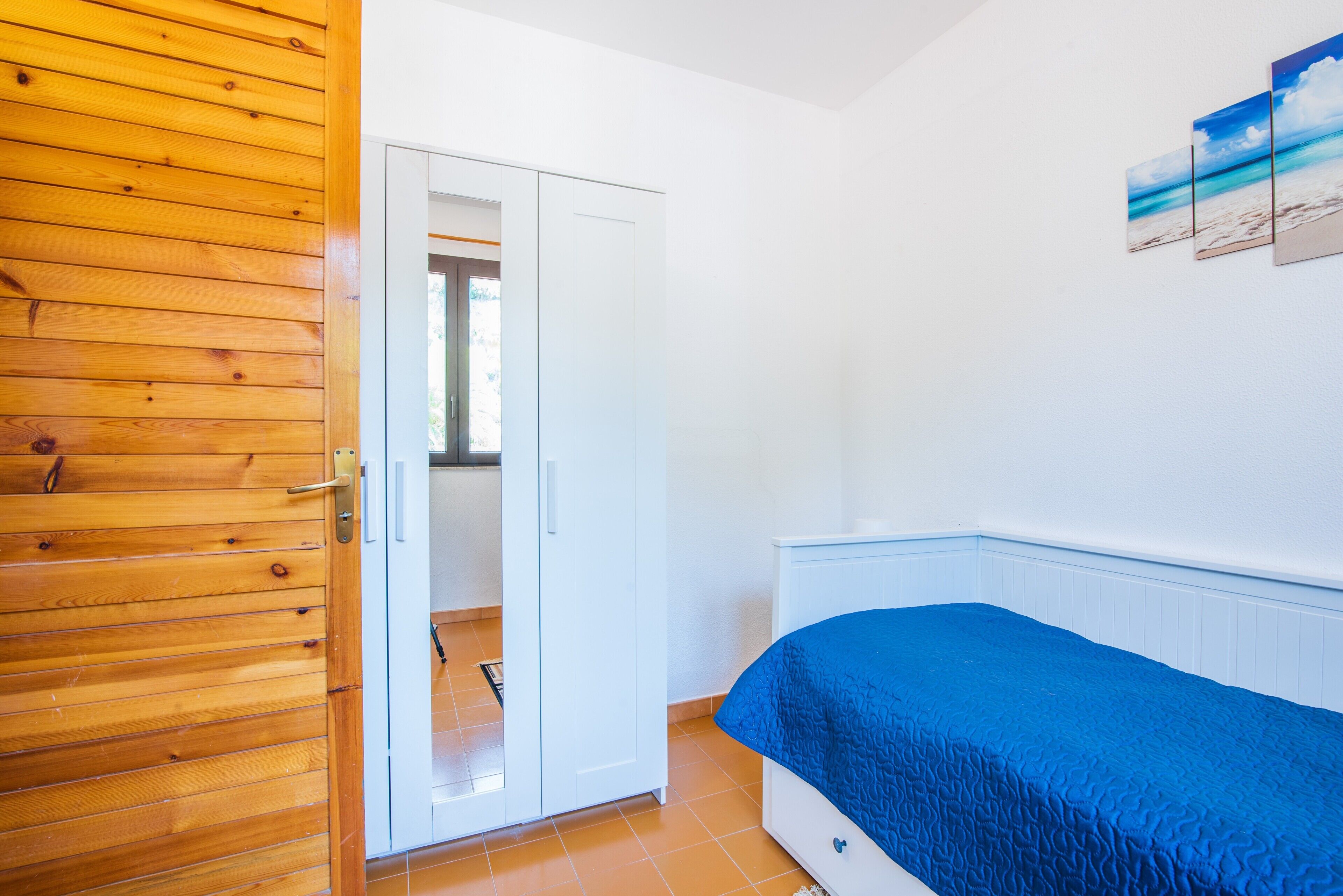 A bedroom with a wooden door, a white wardrobe, a bed with a blue cover, and a wall-mounted picture.