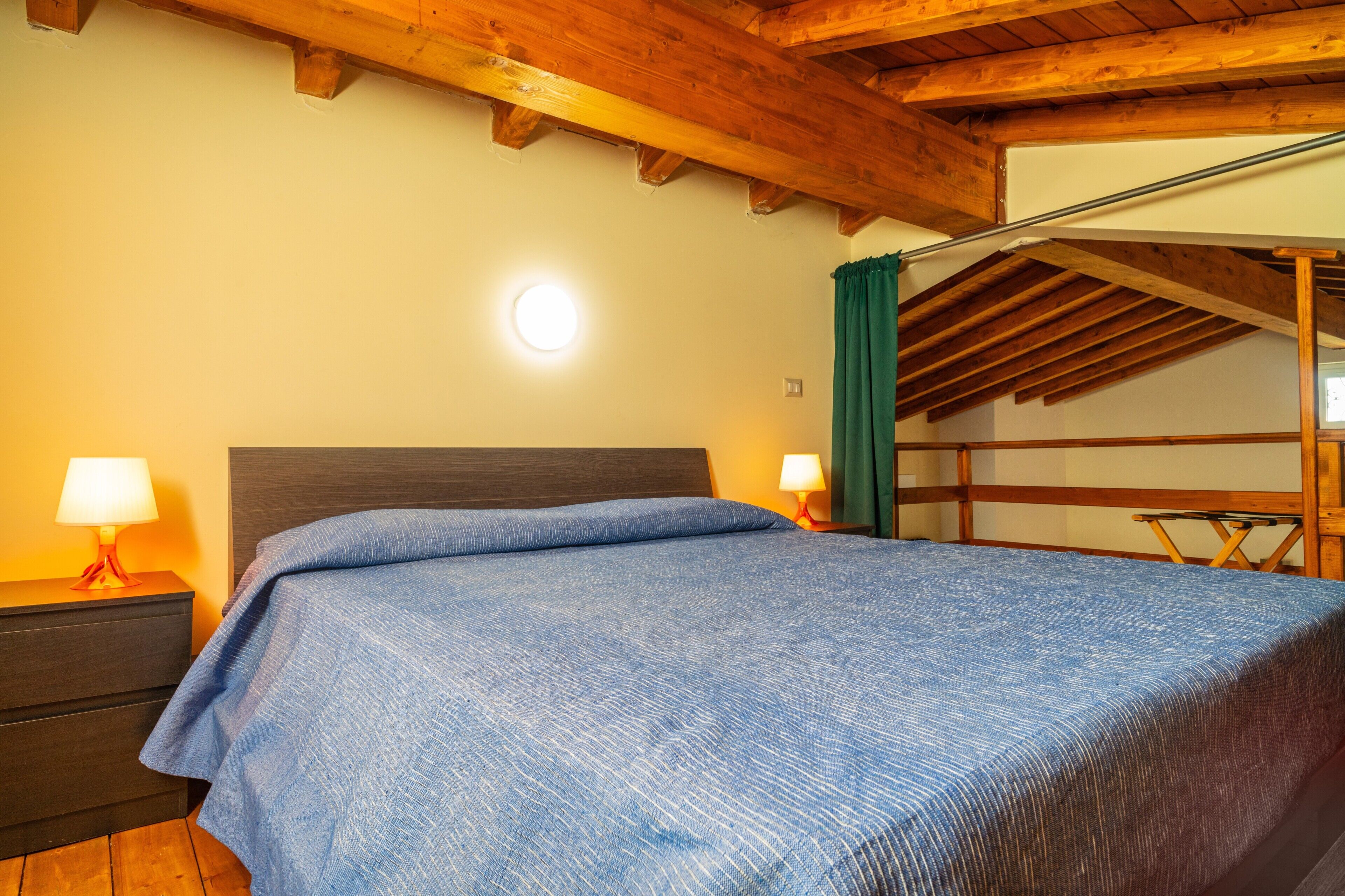 Ein Schlafzimmer mit Holzboden, einem Bett mit blauer Bettdecke, einem Nachttisch mit Lampe und einem Fenster mit Vorhängen.
