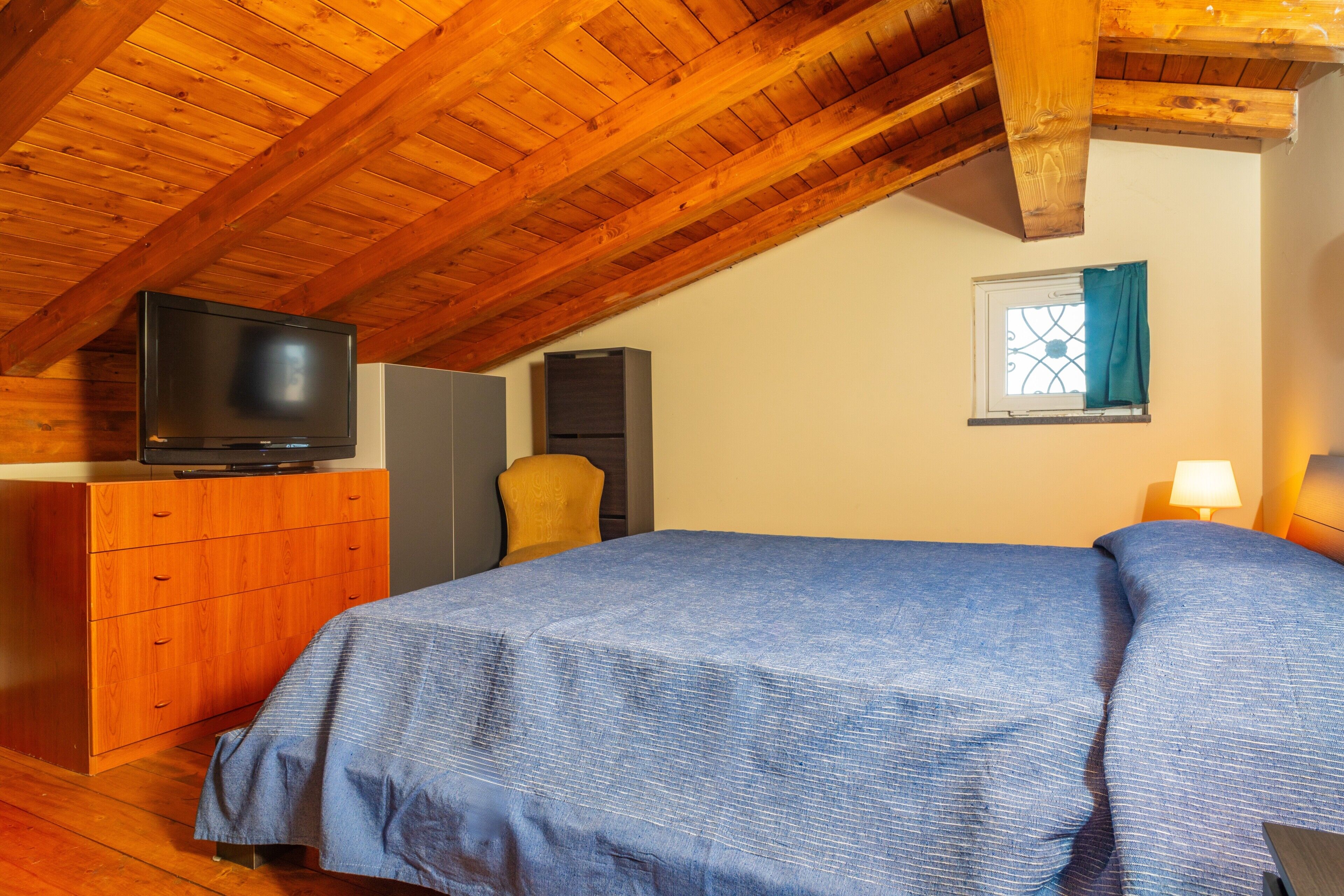 Une chambre avec un plafond en bois, un lit recouvert d’une courtepointe bleue, un téléviseur posé sur une commode en bois, une chaise et une fenêtre avec un rideau.