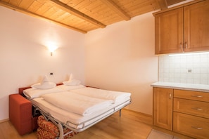 A bedroom with a bed, a small red couch, wooden cabinets, and a tiled backsplash in the adjacent kitchen.