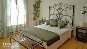 Un dormitori amb estructura de llit metàl·lica, una finestra gran amb cortines lleugeres, una taula de nit amb una làmpada, una planta de bambú alta i un capçal decoratiu metàl·lic.