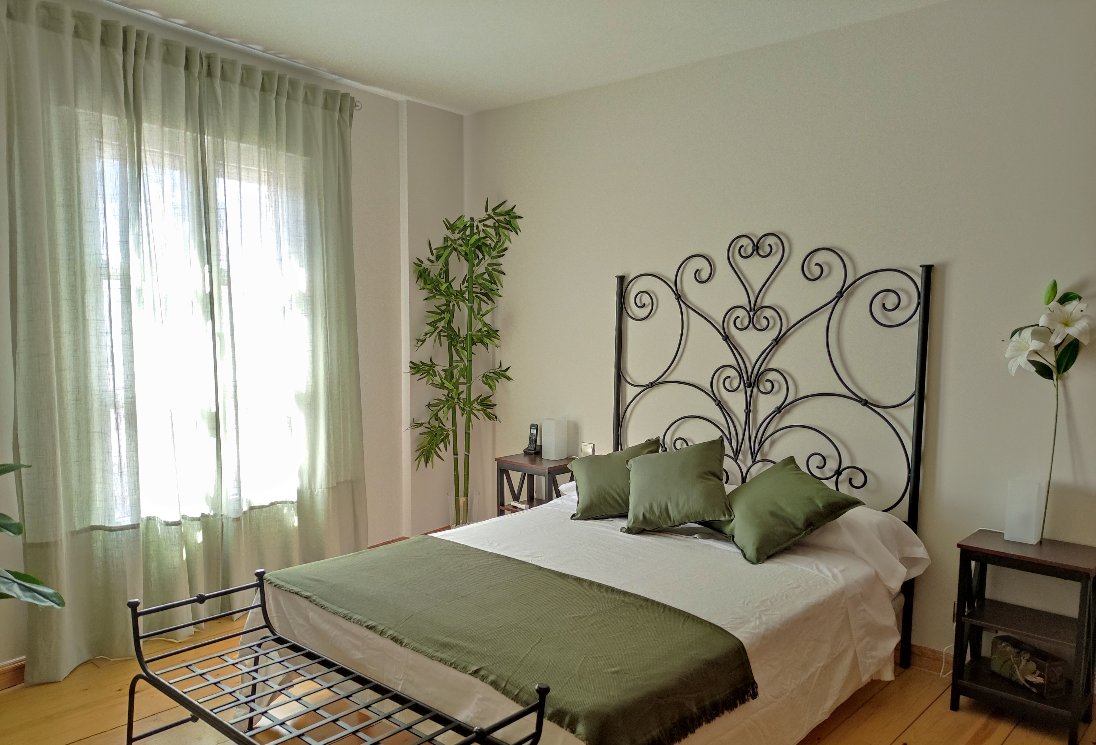 Een slaapkamer met een metalen bedframe, een groot raam met dunne gordijnen, een nachtkastje met een lamp, een hoge bamboeplant en een decoratief metalen hoofdbord.