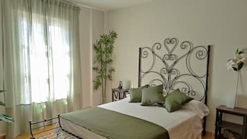 Een slaapkamer met een metalen bedframe, een groot raam met dunne gordijnen, een nachtkastje met een lamp, een hoge bamboeplant en een decoratief metalen hoofdbord.
