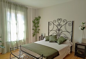 A bedroom with a metal bed frame, a large window with sheer curtains, a bedside table with a lamp, a tall bamboo plant, and a decorative metal headboard.