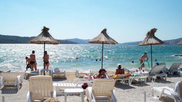 Una playa con sillas de playa blancas, sombrillas de paja y gente nadando y relajándose.