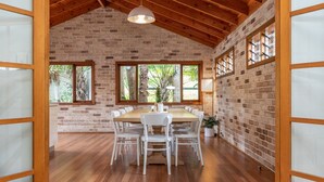 A dining area with a brick wall, wooden ceiling beams, and large windows. - Ponderosa, a gorgeous and spacious five bedroom beach house (Blueys Beach)