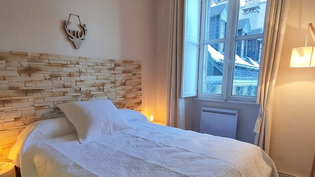 A neatly made bed with a white bedspread and pillows, a stone-patterned headboard, a bedside lamp, and a window with a view of buildings. - 4-person Apartment near Cable Cars in Cauterets (Cauterets)