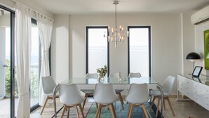 Un espace de salle à manger moderne avec une table en verre, des chaises blanches, un lustre et une vue dégagée grâce à de grandes fenêtres.