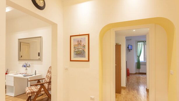 A room with a desk, chair, clock, framed picture, and a hallway leading to another room. - Appartamento Vaticano per 6 persone (Rome)