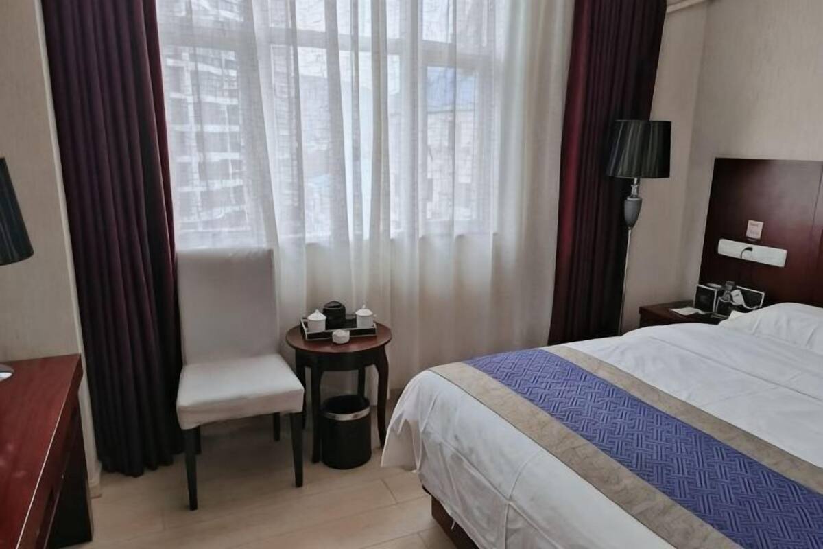 Ein Hotelzimmer mit Bett, Schreibtisch, Stuhl, kleinem Tisch, Lampe und einem Fenster mit Vorhängen.