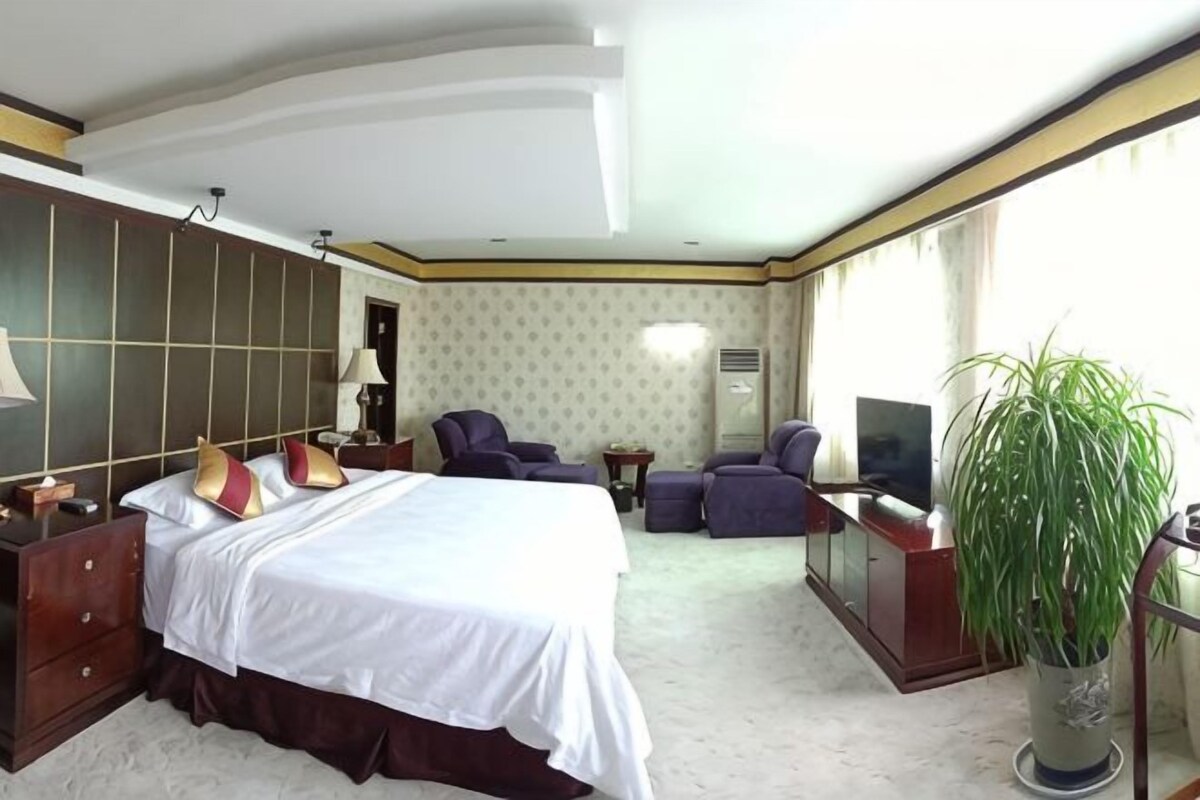 A hotel room with a large bed, a seating area with a sofa and armchairs, a television, a potted plant, and a large window.
