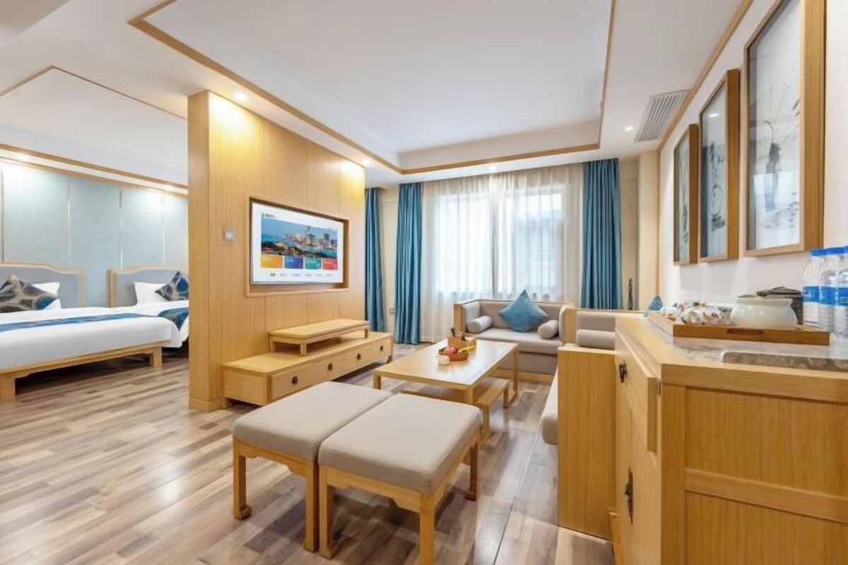 A modern hotel room with a bed, sofa, dining table, bench, and kitchenette.