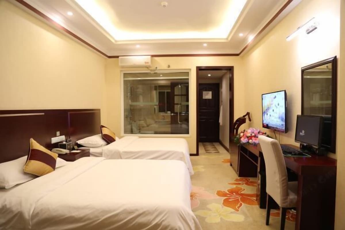 Habitación de hotel con dos camas, un escritorio con computadora, televisión y baño visible a través de una puerta de vidrio.