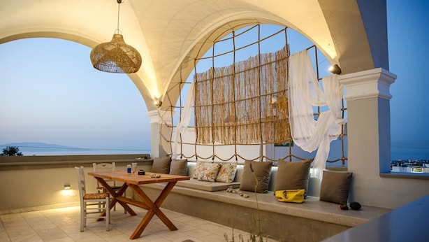 A cozy balcony with a wooden table and chairs, a hanging light fixture, and a view of the sea.