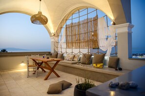 A cozy balcony with a wooden table and chairs, a hanging light fixture, and a view of the sea. - Central Oia Hostel (Santorini)