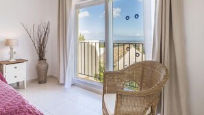 Een balkon met uitzicht op de stad, een rieten stoel, een nachtkastje met een lamp en een vaas met takken.
