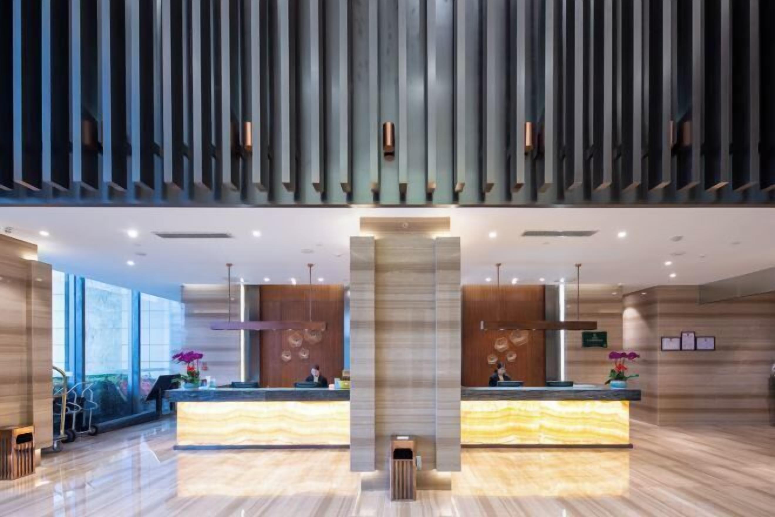A modern hotel lobby with two reception desks, large windows, and a high ceiling with vertical panels.
