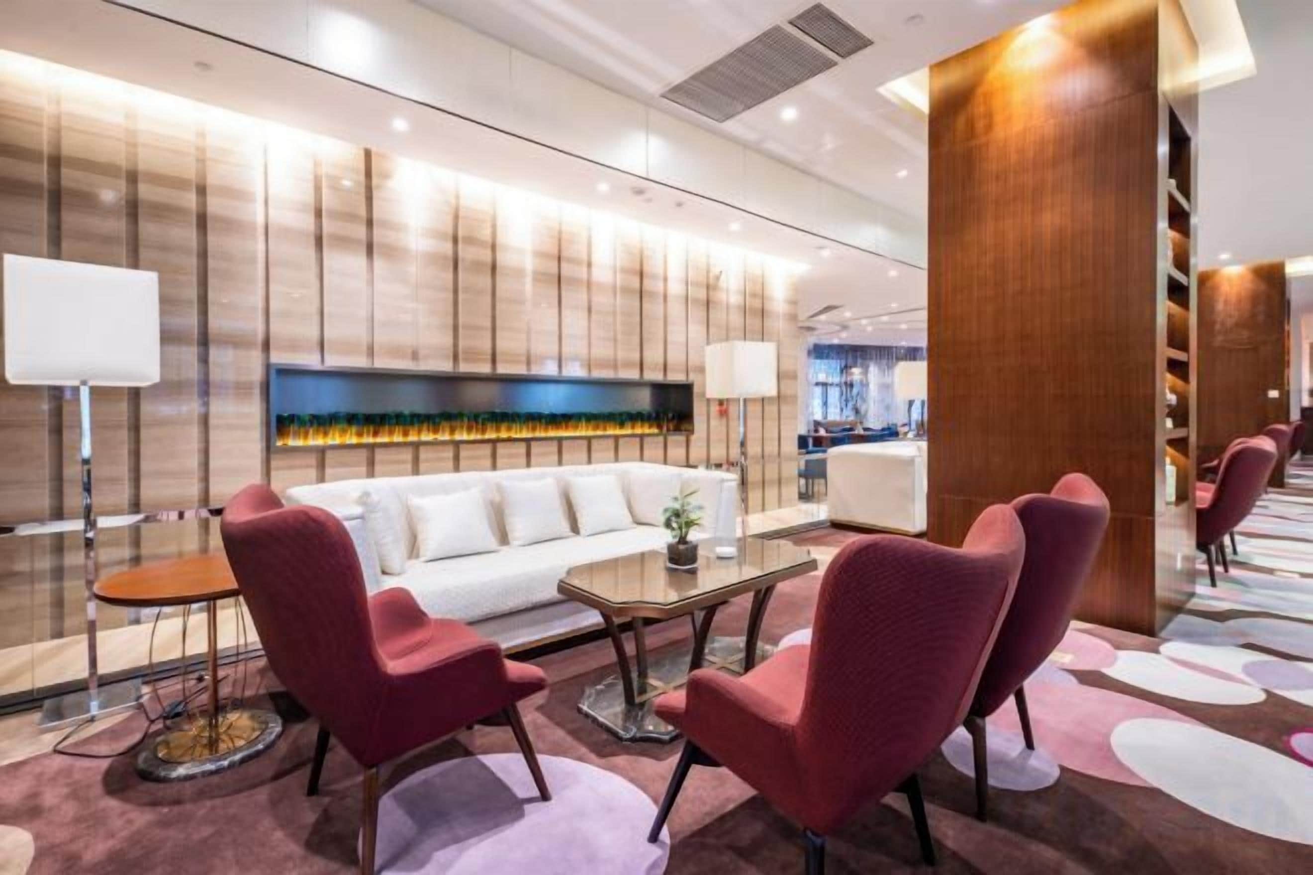 A modern hotel lobby with a sofa, red chairs, a coffee table, and a fireplace.