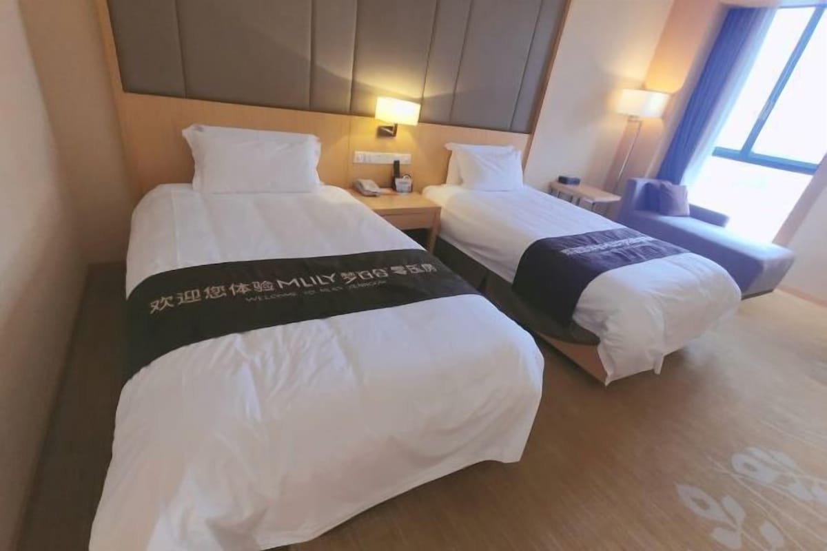 Habitación de hotel con dos camas, cada una con una colcha blanca y una manta negra con texto. Entre las camas, un mesita de noche con una lámpara y un teléfono. El piso es de madera y hay una ventana con cortinas.