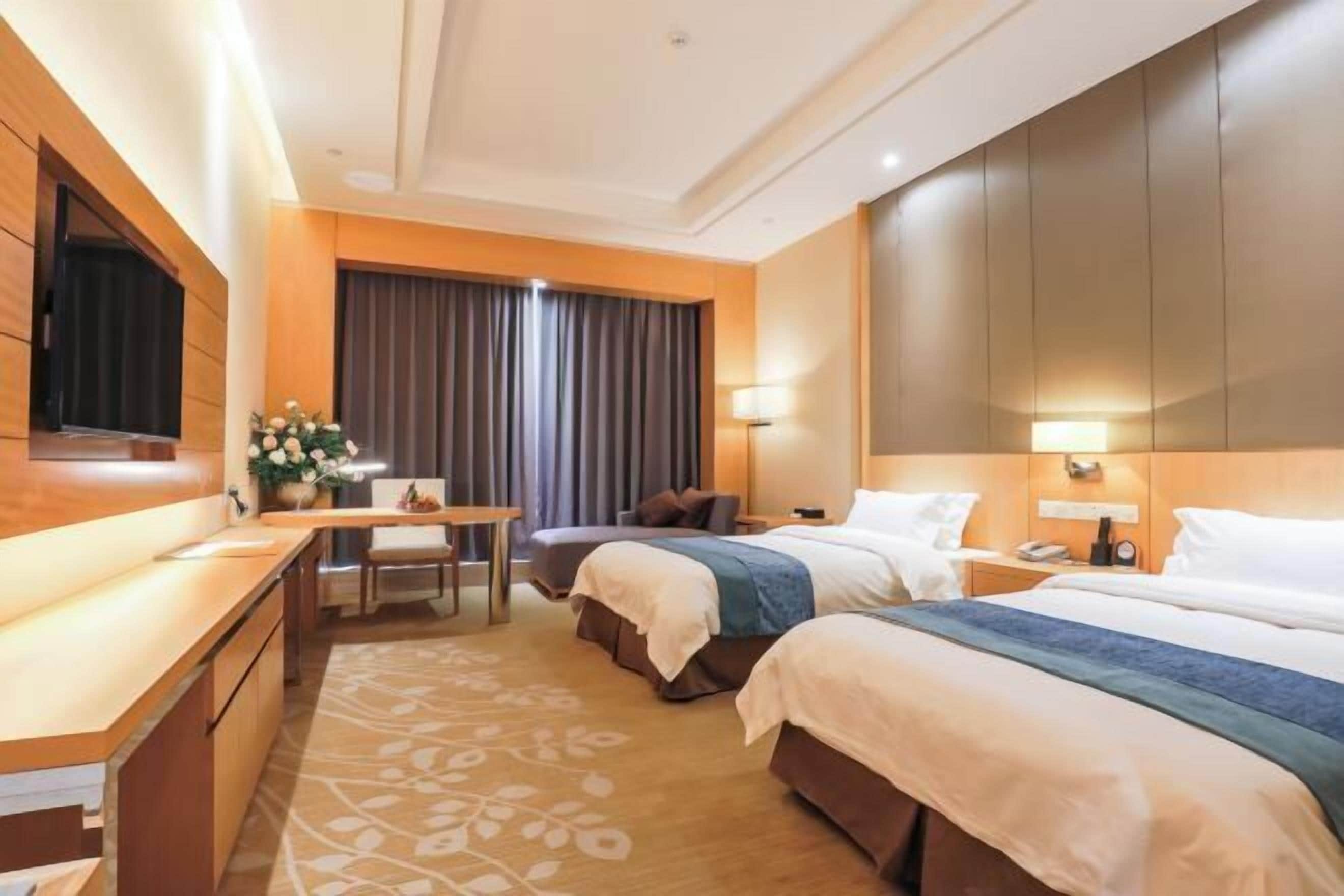 Quarto de hotel com duas camas, uma mesa, televisão e uma janela grande com cortinas.