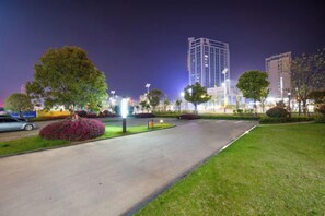 A well-lit urban street at night with a tall building in the background, a clear road, and green landscaping. - Golden Sun Hotel (Lai'an)