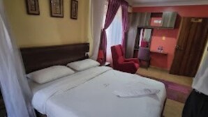 Une chambre d’hôtel avec un grand lit, un bureau et une chaise. Les murs sont ornés de tableaux encadrés et il y a une fenêtre avec des rideaux.