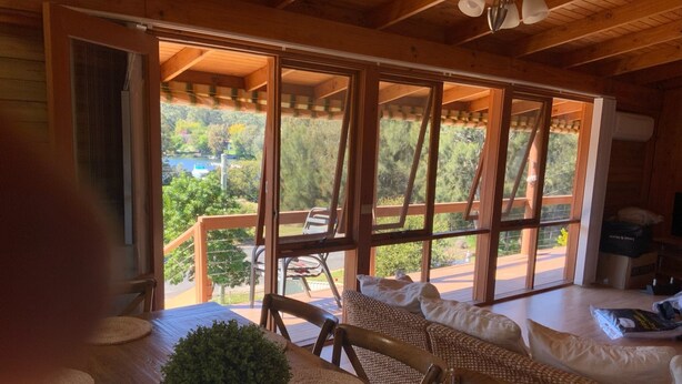 Un intérieur en bois comprenant une table à manger, des chaises et un canapé. De grandes fenêtres offrent une vue sur la verdure et un bâtiment situé à l’extérieur.