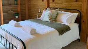 Une chambre dans une cabane en bois, avec un lit en métal, du linge de lit blanc et un ventilateur de plafond.
