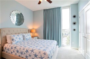 A bedroom with a bed, a nightstand, a ceiling fan, a window with curtains, and a view of the outside. - Lagoon Tower 302, 3/3 | Private Beach Access (Gulf Shores)