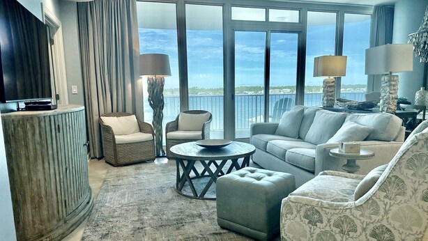 Amplia sala de estar con un ventanal que ofrece vistas al mar, un televisor sobre un mueble de madera y una zona de estar acogedora con un otoman estampado.