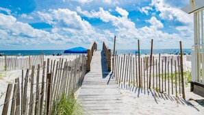 Una pasarela de madera que lleva a una playa con cielo despejado y una montaña distante.