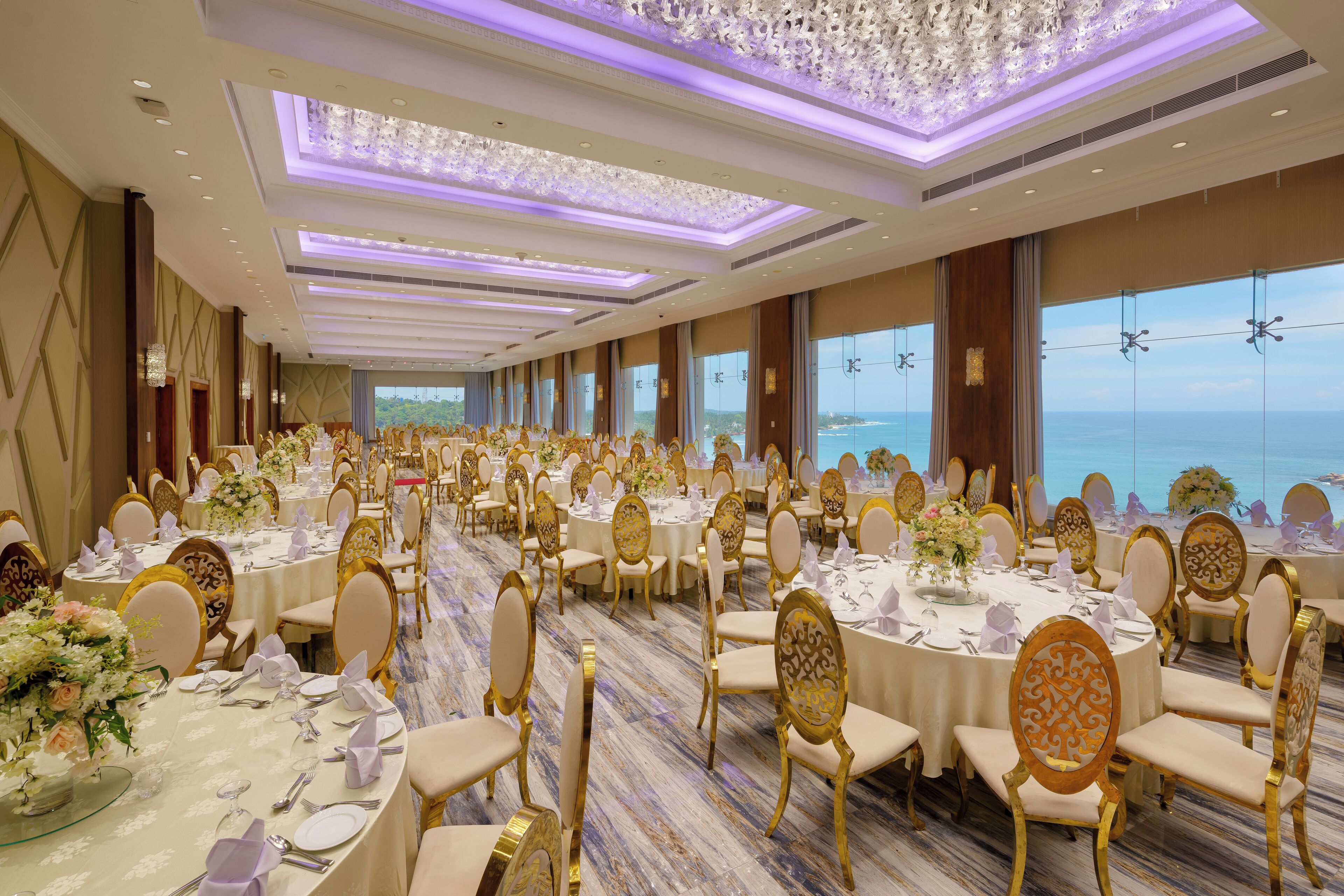 Un salón de banquetes con mesas redondas, sillas doradas y vistas al mar.