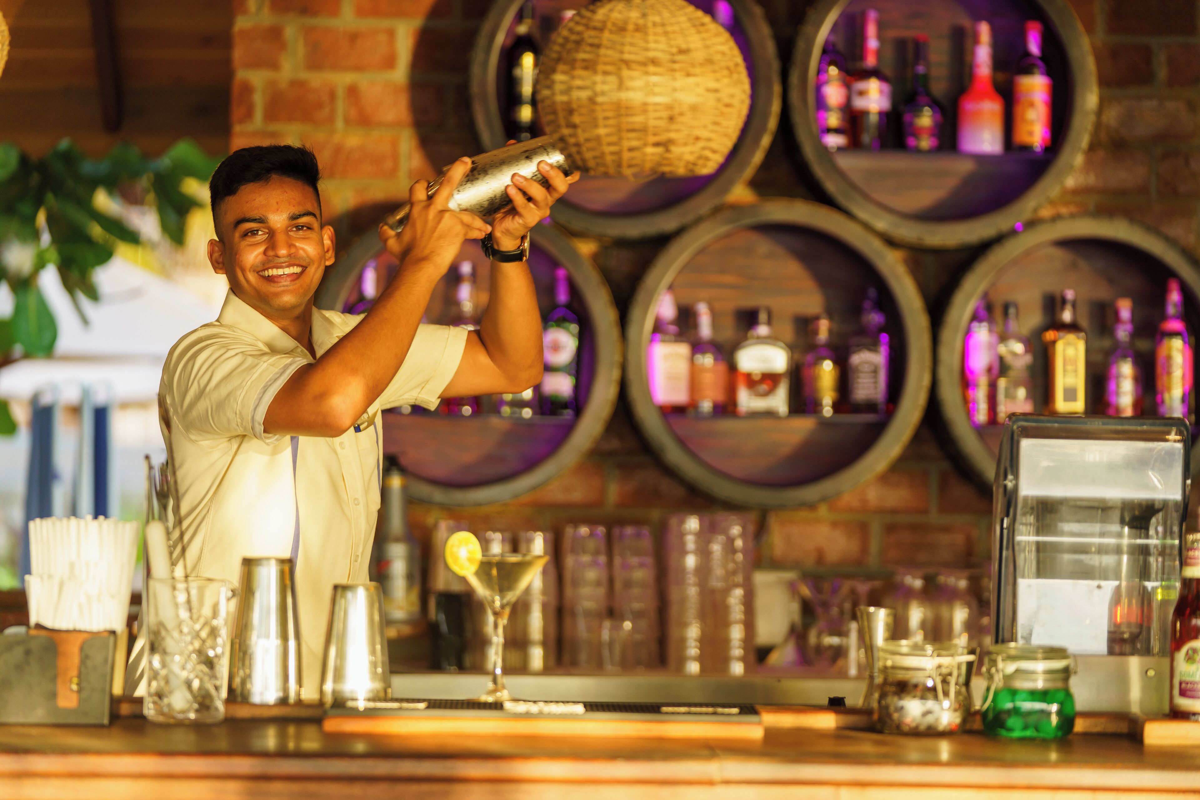 A bartender preparing a drink at a bar with wine barrels in the background.