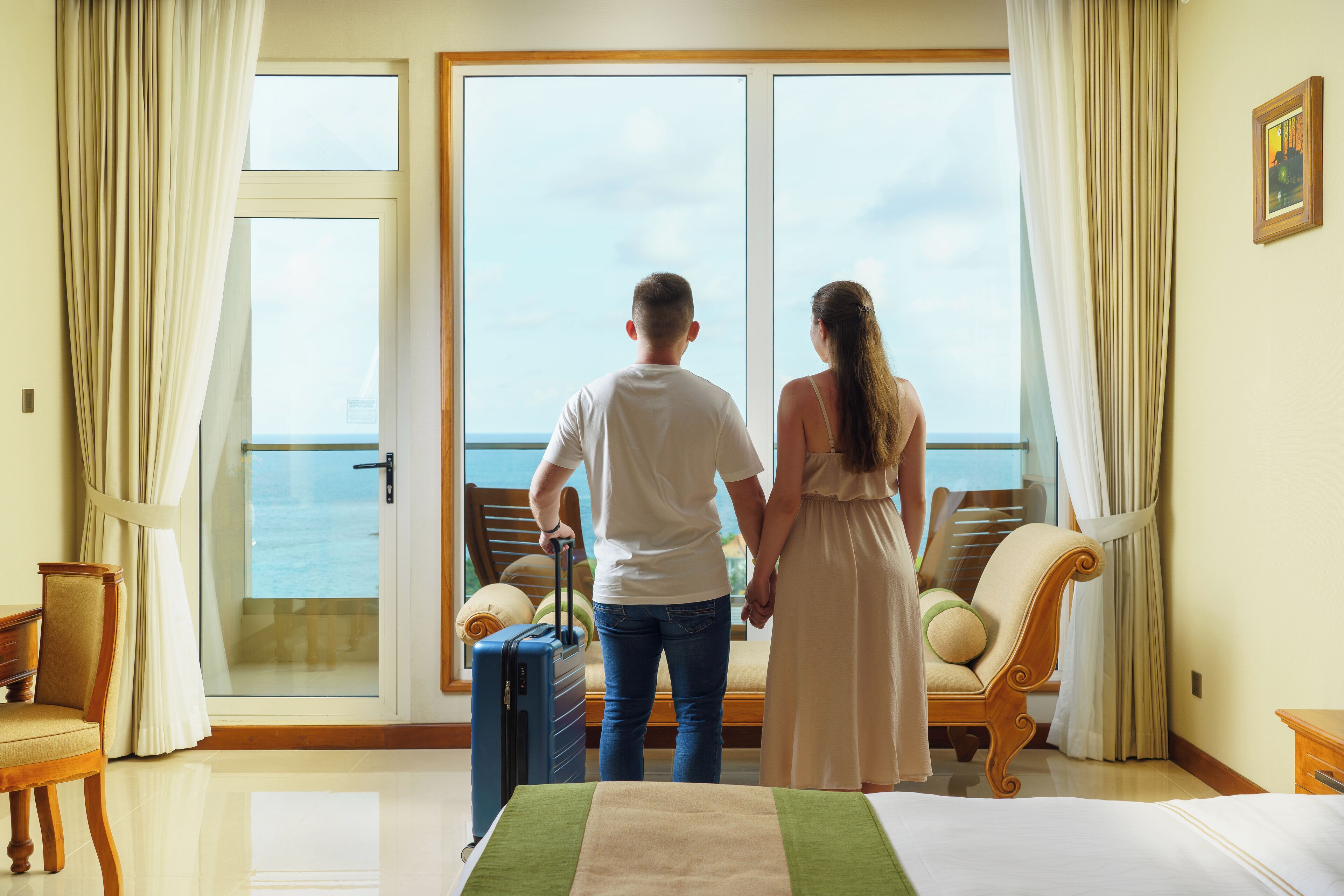 Een koppel staat bij een groot raam met uitzicht op zee, met een bed en een koffer op de voorgrond.
