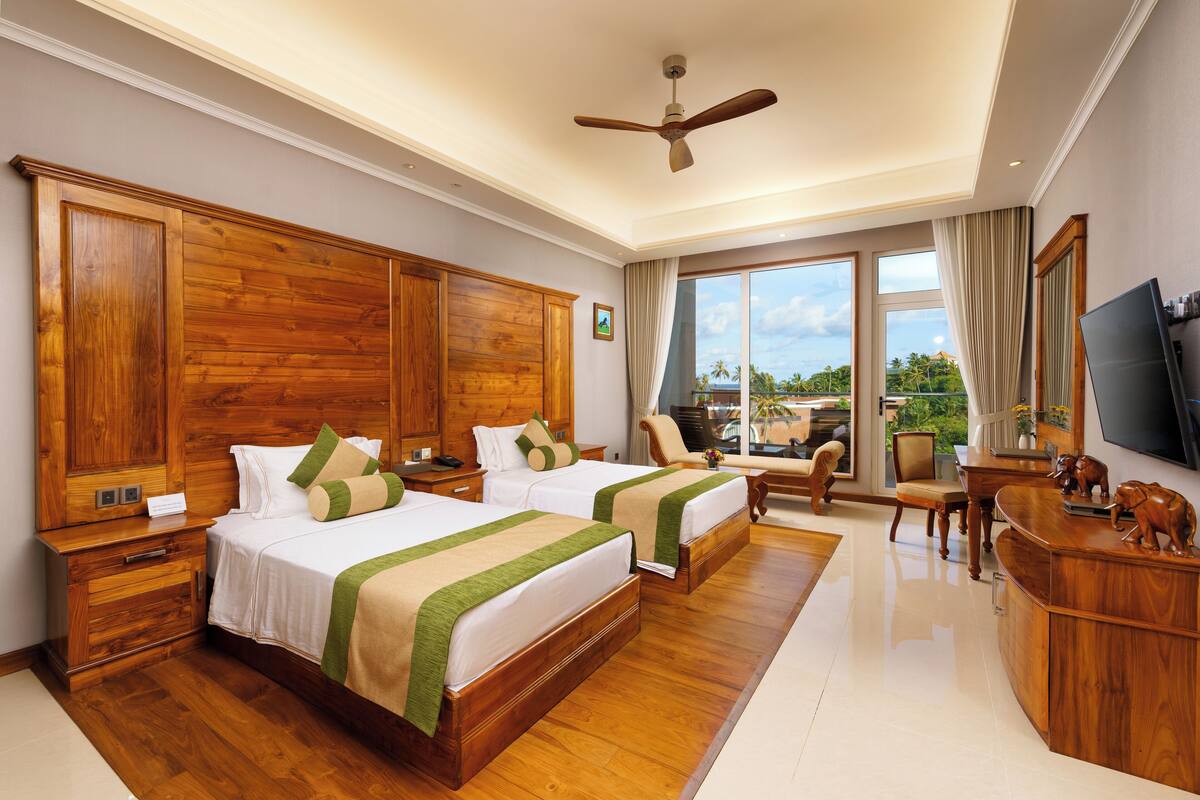 A hotel room with two beds, wooden furniture, a ceiling fan, a TV, and a view of the outdoors.
