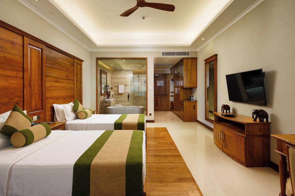 A hotel room with two beds, wooden headboards, a flat-screen TV, a wooden cabinet, and a bathroom visible through an open door.