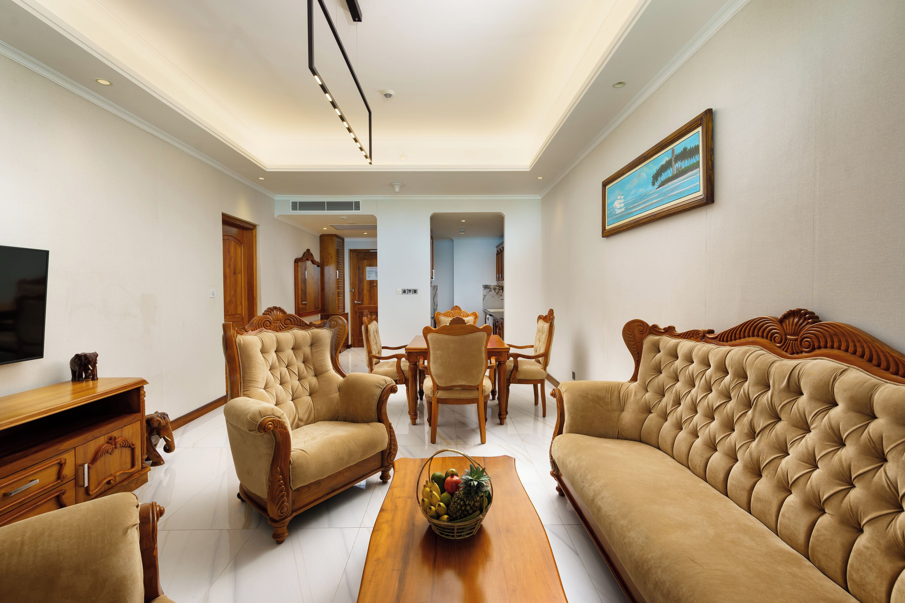 A spacious living room with a large sofa, two armchairs, a wooden coffee table with fruit, a dining area with a table and chairs, and a television on the wall.