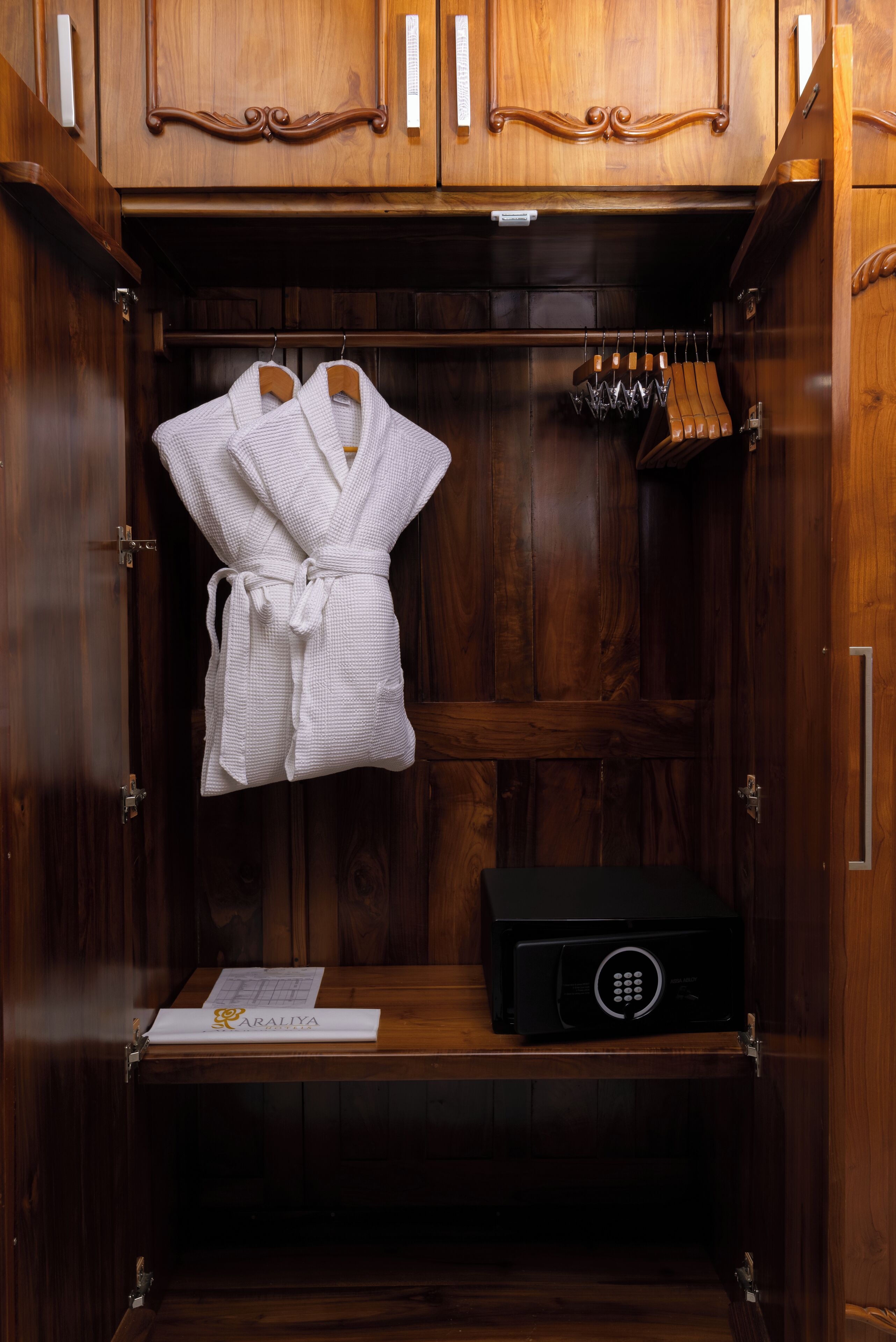 A wooden wardrobe with a robe hanging inside, a black safe, and a set of wine glasses on the top shelf.