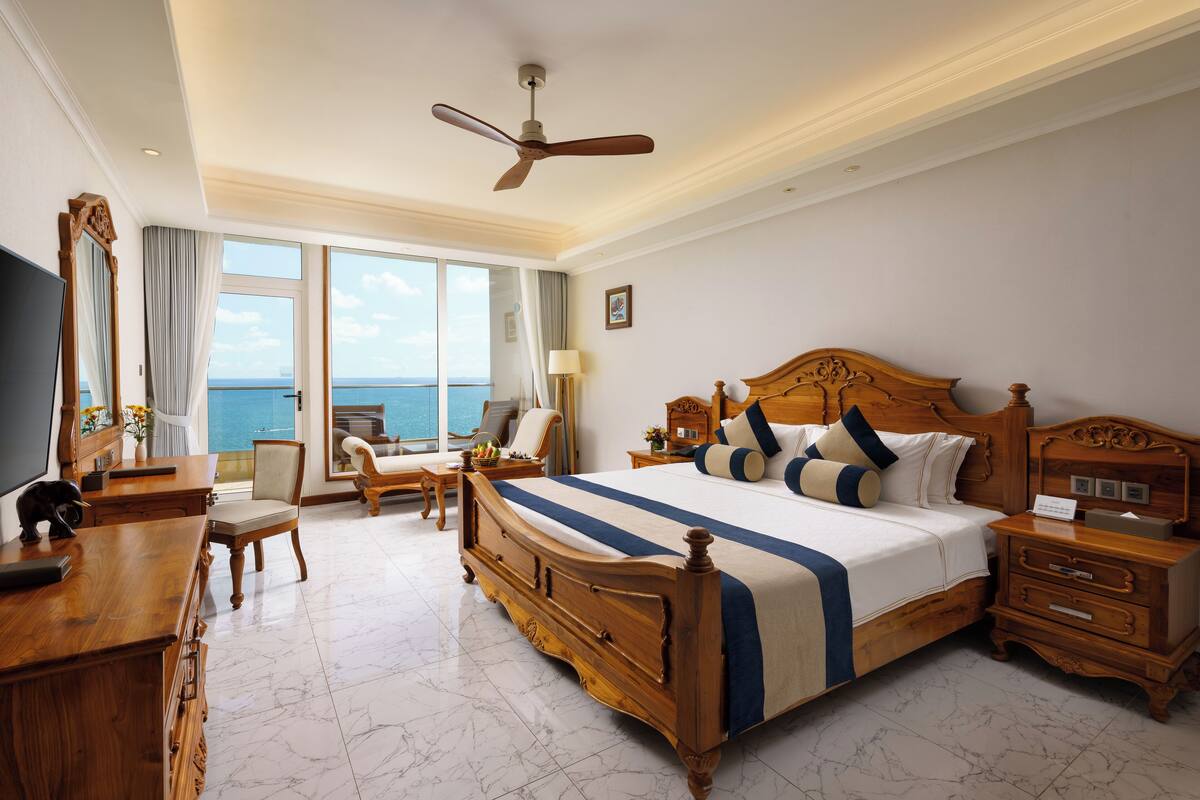 A spacious hotel room with a large bed, wooden furniture, a desk, and a view of the sea.
