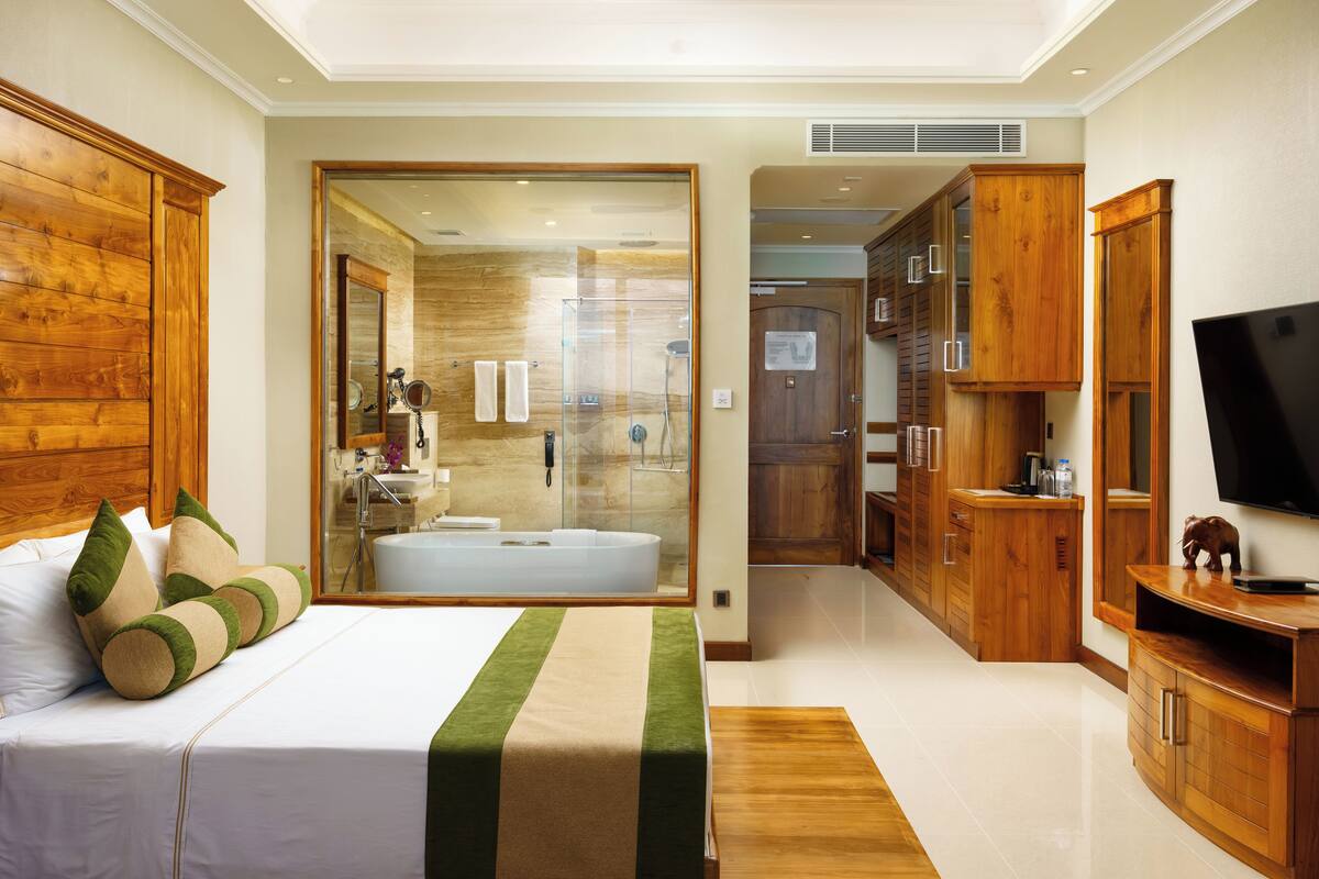 A modern hotel room with a large bed, wooden furniture, a TV, and a bathroom with a bathtub.