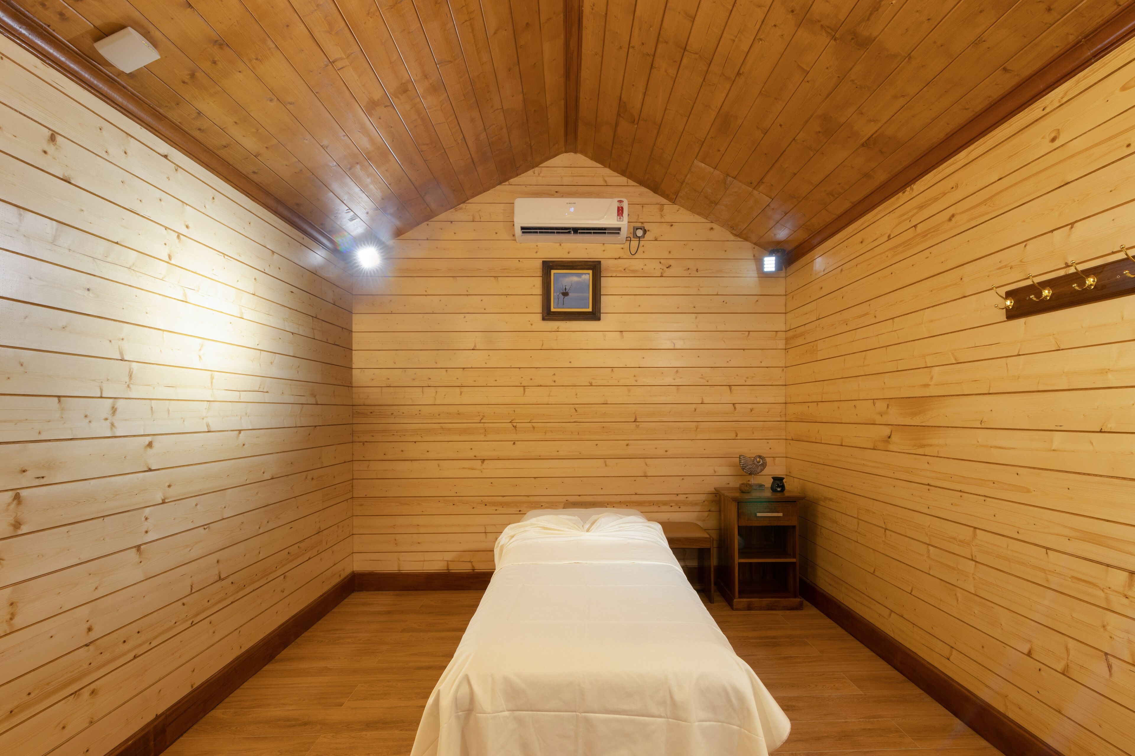A small, wooden-paneled room with a single bed, a small table, and a wall-mounted air conditioning unit.
