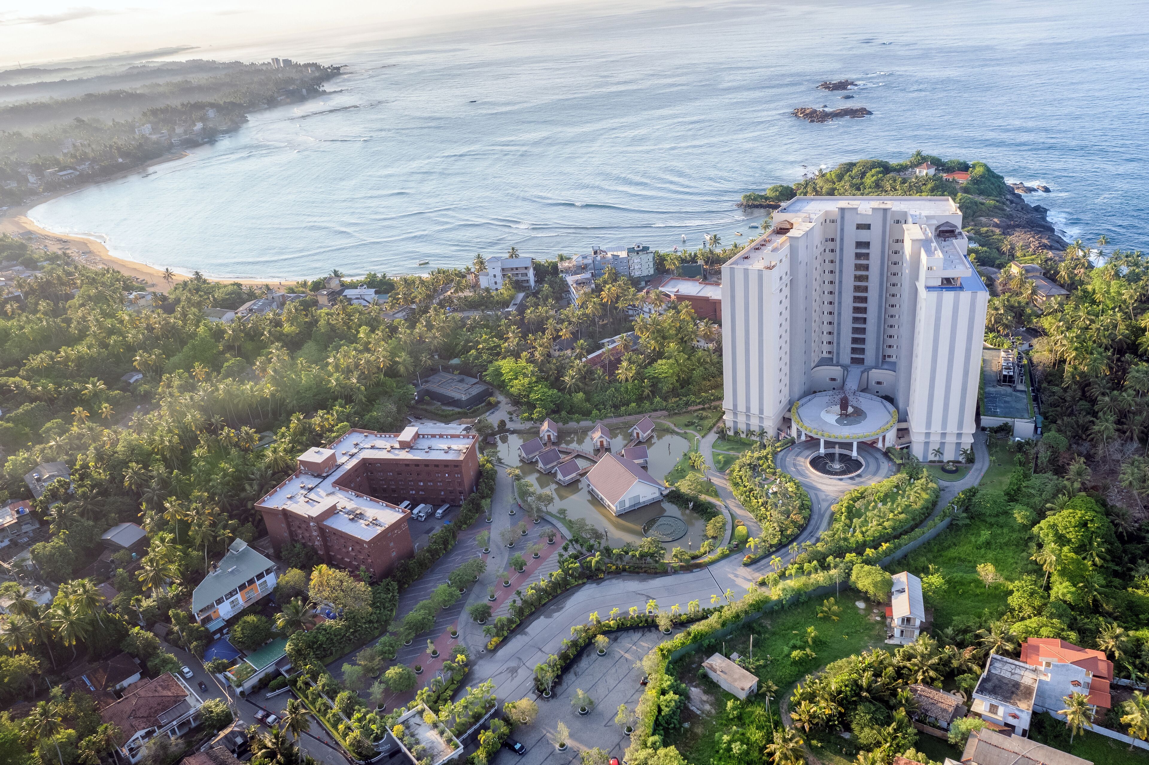 Vue aérienne d’un hôtel situé sur la côte, avec un bâtiment central, une plage et une végétation luxuriante tout autour.