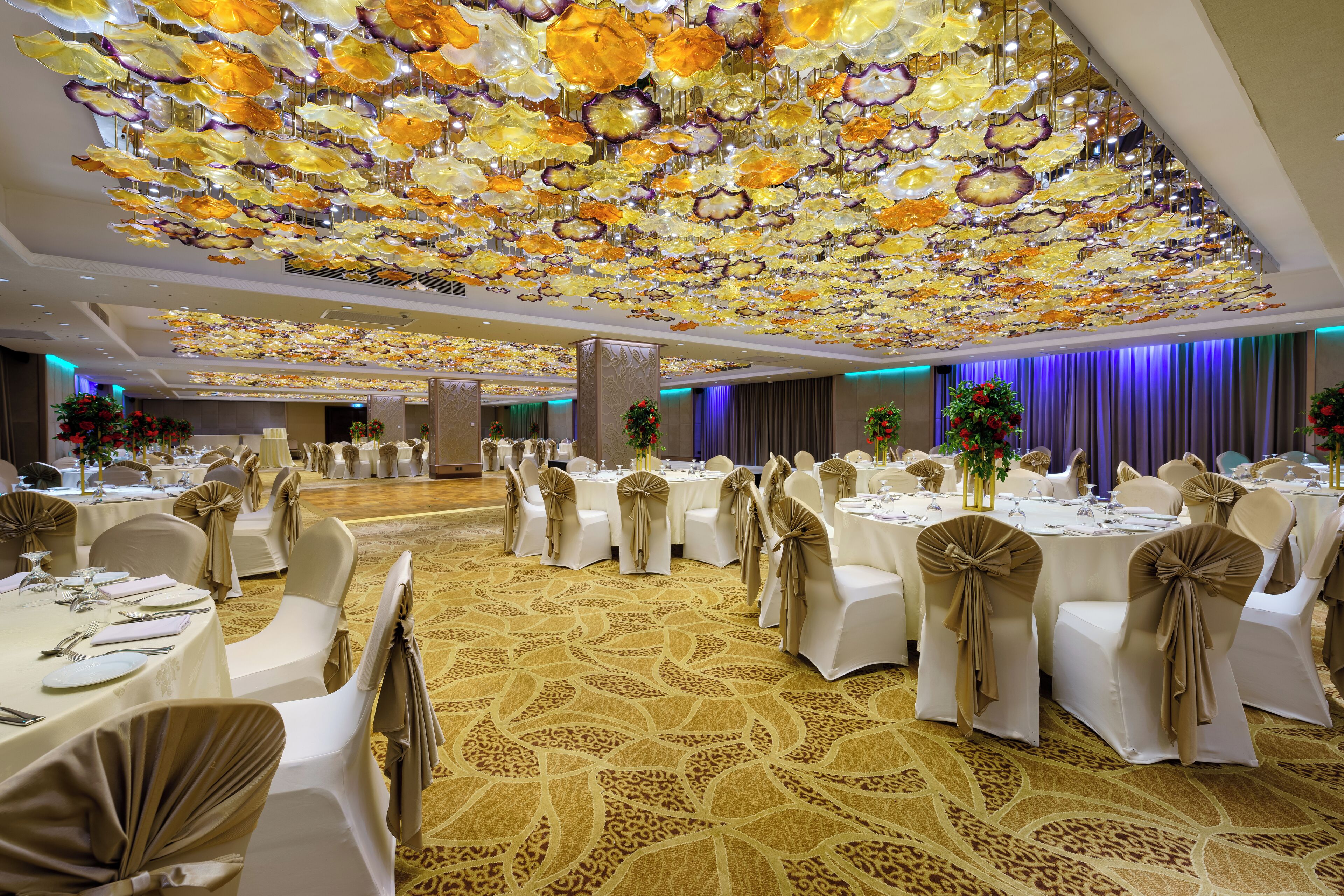 A banquet hall with round tables set for dining, white chairs with beige sashes, and a ceiling adorned with floral decorations.