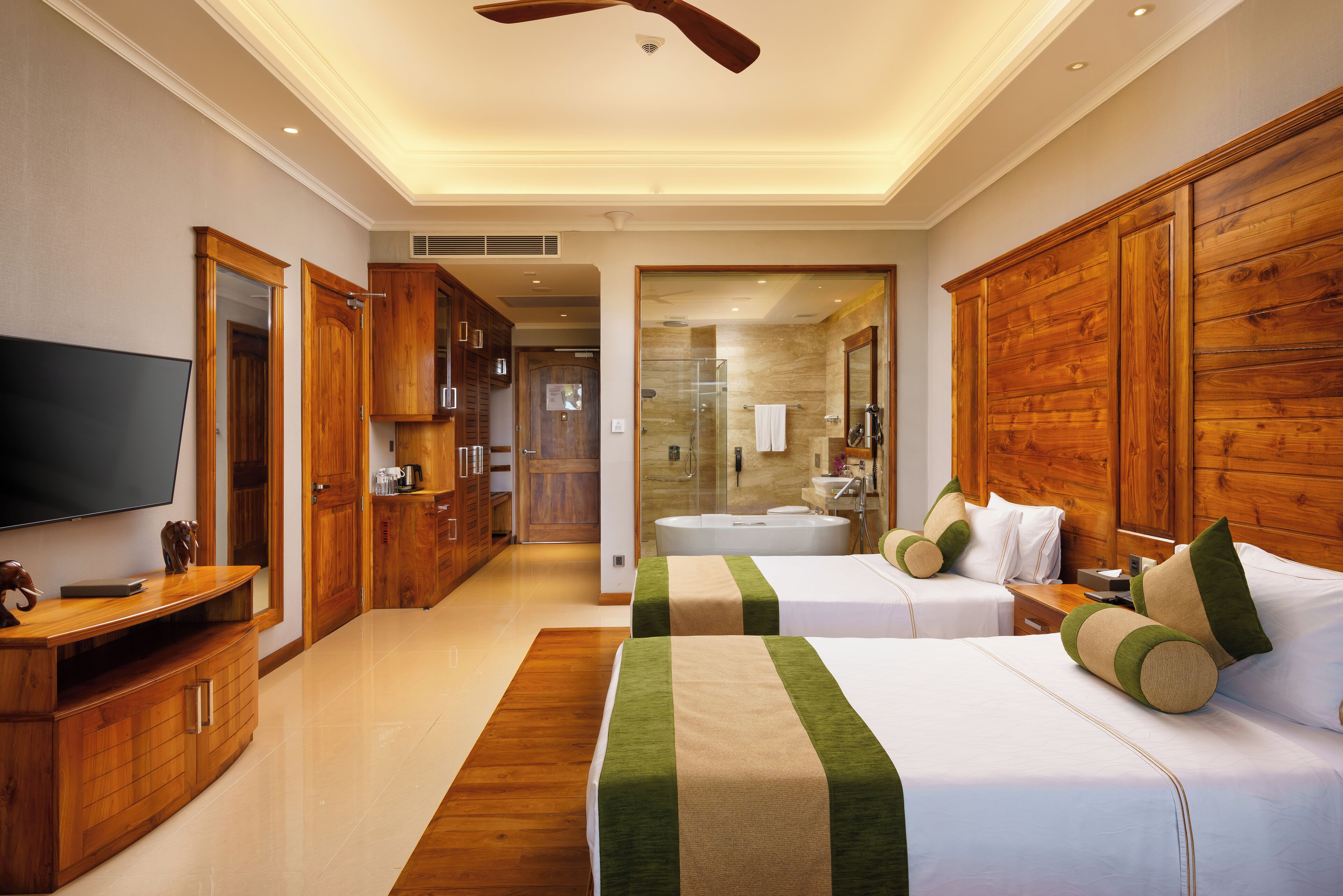 Habitación de hotel con televisión, muebles de madera, cama con ropa de cama verde y beige, y baño con bañera y ducha.