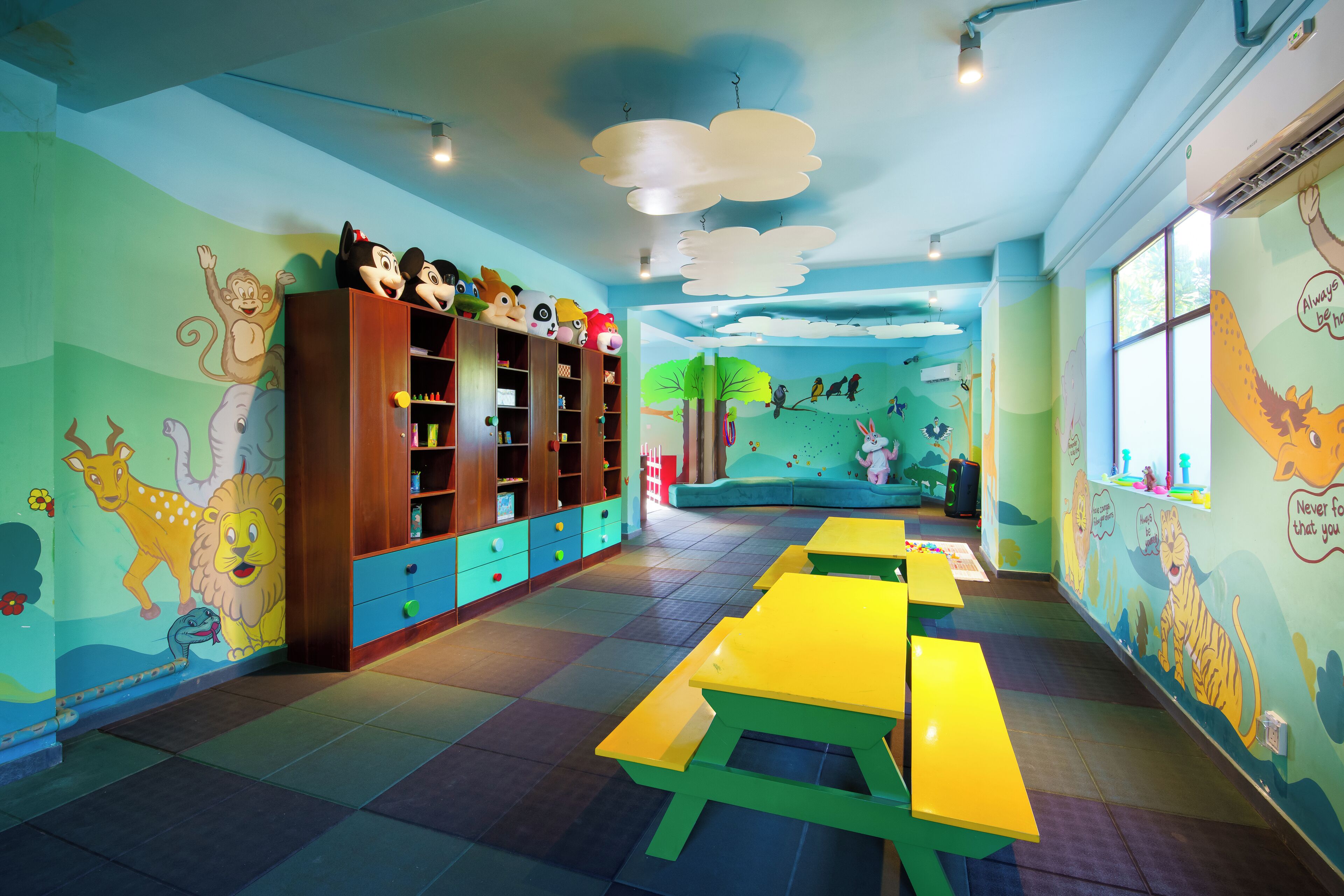 A children's playroom with colorful murals, a long table, and a bookshelf filled with toys.