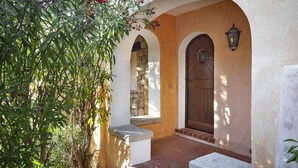 Un portail en arc surmonté d’un mur de pierre et d’une porte en bois, qui mène à un patio abrité avec un banc en pierre et un pot de fleurs.