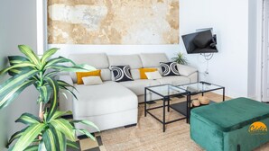 A living room with a sofa, a coffee table, a green ottoman, a television mounted on the wall, and a large plant.